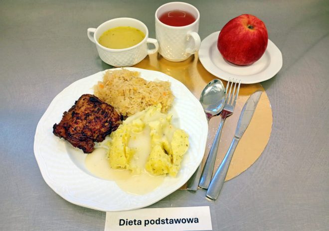 Obiad „Dieta podstawowa” na metalowym blacie: na białym talerzu porcja pieczonego/smażonego mięsa, ziemniaki z koperkiem polane jasnym sosem oraz duszona kapusta. W tle stoją miseczka z jasną zupą i kubek z czerwonym napojem, a obok na spodeczku leży czerwone jabłko. Po prawej stronie widoczne sztućce oraz kartka z napisem „Dieta podstawowa”