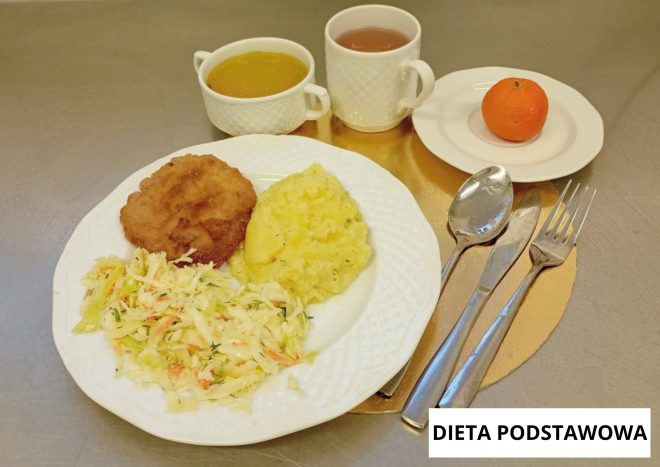 Obiad w diecie podstawowej: kotlet panierowany, ziemniaki purée i surówka z kapusty na białym talerzu; obok miseczka zupy, kubek herbaty, mandarynka na osobnym talerzyku oraz sztućce