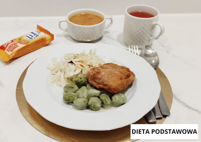 Obiad w diecie podstawowej podany na białym talerzu. Na talerzu znajduje się kotlet mielony, kluski oraz surówka z białej kapusty. W tle widoczna jest miseczka z zupą oraz kubek z herbatą. Zestaw ułożony na jasnym blacie, z oznaczeniem „Dieta podstawowa”