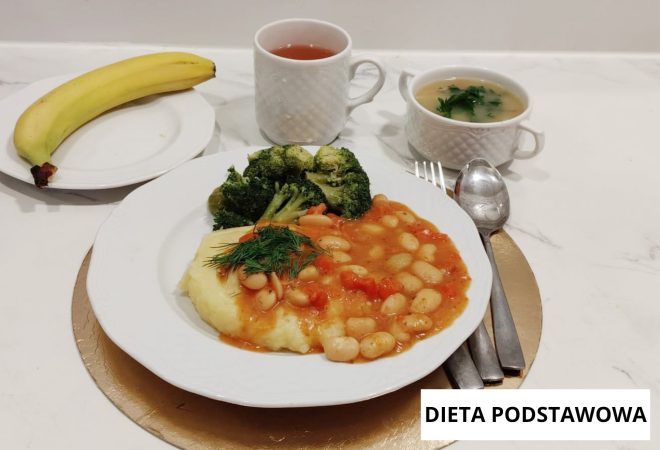 Obiad w diecie podstawowej: fasolka w sosie pomidorowym podana z puree ziemniaczanym, gotowanymi brokułami i koperkiem. Obok talerza banan, zupa jarzynowa oraz herbata. Na zdjęciu widoczny napis „Dieta podstawowa”