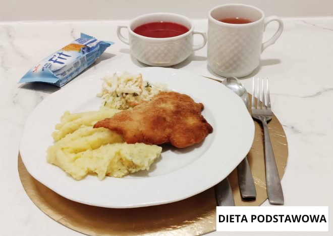 Obiad w diecie podstawowej: panierowany kotlet smażony podany z puree ziemniaczanym i surówką z kapusty. Obok talerza miseczka z czerwonym kompotem oraz kubek z herbatą. Na zdjęciu widoczny napis „Dieta podstawowa”