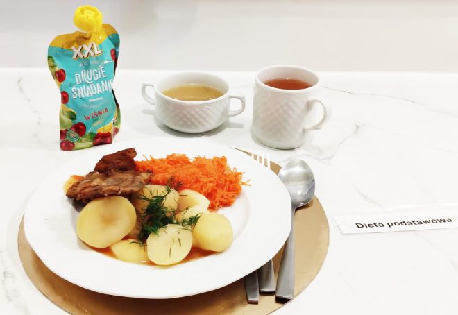 Na białym stole zestaw obiadowy: talerz z porcją mięsa w sosie, gotowanymi ziemniakami posypanymi koperkiem oraz surówką z marchewki. Obok kubek herbaty, filiżanka jasnego napoju oraz mus owocowy „Drugie śniadanie” o smaku wiśni. Przy talerzu sztućce i karteczka z napisem „Dieta podstawowa”