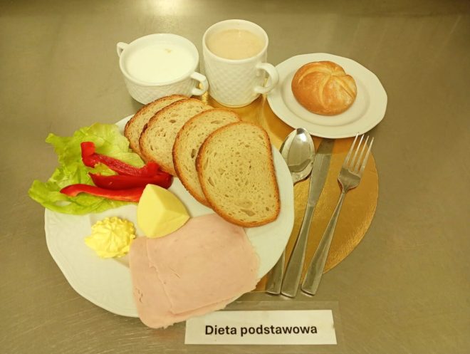 Śniadanie w diecie podstawowej: kanapki z pieczywa, obok masło, ser żółty, wędlina oraz papryka i sałata. Obok w filiżance jest zupa mleczna oraz kawa w kubku i bułka kajzerka na osobnym talerzyku.