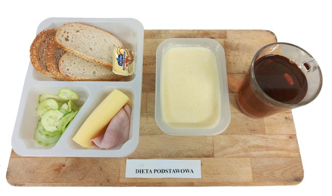 Śniadanie szpitalne – dieta podstawowa: pieczywo pszenno-żytnie z masłem, plaster sera żółtego i wędliny drobiowej, świeże ogórki, zupa mleczna oraz herbata. Posiłek zaprezentowany w pojemnikach jednorazowych na drewnianej desce