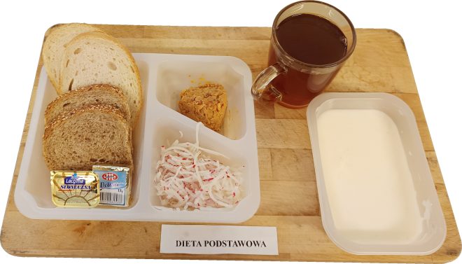 Śniadanie w diecie podstawowej: kromki pieczywa pszennego i razowego z masłem, pasta kanapkowa, surówka z białej rzodkwi, herbata oraz jogurt naturalny, ułożone na drewnianej desce