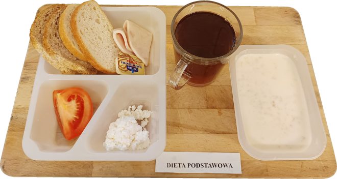 Śniadanie w diecie podstawowej: pieczywo mieszane z wędliną drobiową i masłem, twarożek, plaster pomidora, herbata oraz zupa mleczna, podane w porcjach na drewnianej desce