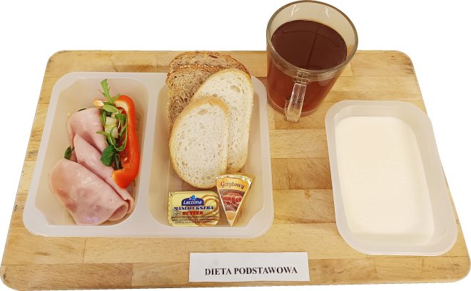 Na drewnianej desce ułożono zestaw śniadaniowy w białej, dwudzielnej tacce. W lewej części znajdują się plastry wędliny zwinięte w roladki, obok pasek czerwonej papryki oraz listki zieleniny. W prawej części leżą kromki pieczywa: jasne i ciemniejsze. Na dole tacki widać porcję masła w złotym opakowaniu oraz trójkątny serek topiony (opis „Grzybowy”). U góry po prawej stoi szklany kubek z ciemną herbatą. Po prawej stronie deski znajduje się dodatkowy pusty biały pojemnik. Na dole kadru widoczna kartka z napisem „DIETA PODSTAWOWA”