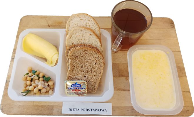 Śniadanie na drewnianej desce: biała tacka z trzema kromkami chleba, zrolowanym plastrem żółtego sera, porcją sałatki z ciecierzycy z zieleniną oraz małą porcją masła w opakowaniu. Obok stoi szklanka z herbatą, a po prawej w pojemniku znajduje się jasna zupa mleczna/owsianka z żółtymi drobinkami. Na dole widoczna kartka z napisem „DIETA PODSTAWOWA”
