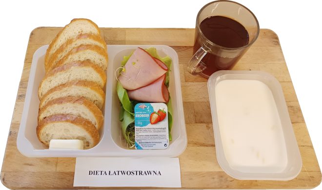 Śniadanie w diecie łatwostrawnej: w pojemniku kromki jasnego pieczywa z porcją masła, plastry szynki ułożone na liściach sałaty oraz mały pojemnik dżemu truskawkowego; obok kubek herbaty i pojemnik z mlekiem. Całość ustawiona na drewnianej desce