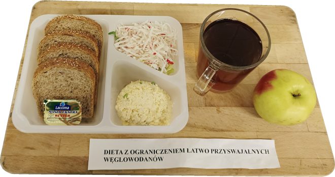 Śniadanie w diecie z ograniczeniem łatwo przyswajalnych węglowodanów podane na drewnianej desce. W plastikowej tacce znajdują się kromki chleba pełnoziarnistego, porcja twarożku oraz surówka z białej rzodkwi. Obok ustawiona jest szklanka herbaty oraz jabłko. Na desce widoczna kartka z napisem „Dieta z ograniczeniem łatwo przyswajalnych węglowodanów”