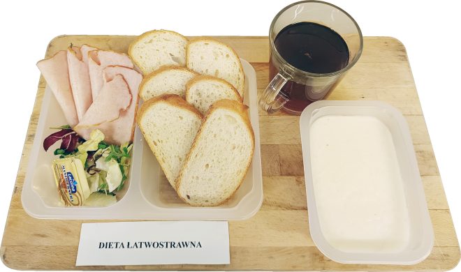 Śniadanie na diecie lekkostrawnej: kromki jasnego pieczywa, wędlina drobiowa, masło, mix sałat, herbata oraz porcja jogurtu naturalnego, ułożone na tackach na drewnianym blacie