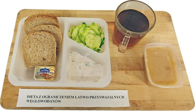 Zestaw śniadaniowy ułożony na drewnianej desce, opisany jako „Dieta z ograniczeniem łatwo przyswajalnych węglowodanów”. W plastikowym pojemniku znajdują się kromki pieczywa razowego, porcje świeżego ogórka pokrojonego w plastry oraz pasta twarogowa z dodatkiem ziół. W osobnej przegródce umieszczono porcję masła w opakowaniu jednorazowym. Po prawej stronie stoi szklany kubek z ciemnym napojem, a obok znajduje się zamknięty pojemnik z jasnym płynem, prawdopodobnie zupą lub napojem. Całość przedstawia kompletny, porcjowany posiłek szpitalny