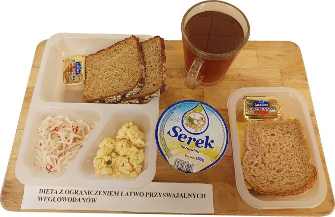 Na drewnianej desce zaprezentowane śniadanie w diecie z ograniczeniem łatwo przyswajalnych węglowodanów. Po lewej stronie tacka z kromkami ciemnego pieczywa oraz porcją masła. Poniżej na tej samej tacce surówka z białej rzodkwi z dodatkiem czerwonej papryki oraz jajecznica. Pośrodku deski opakowanie serka naturalnego. Po prawej stronie tacka z kromkami pieczywa i porcją masła oraz szklany kubek z ciemnym napojem. Na dole deski widoczna etykieta z napisem: ‘Dieta z ograniczeniem łatwo przyswajalnych węglowodanów’