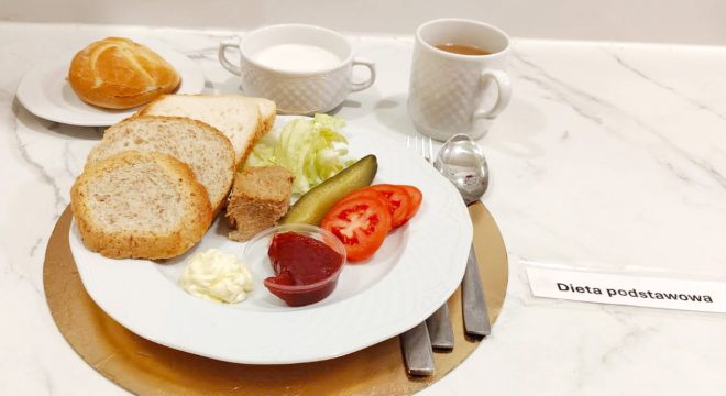 Posiłek diety podstawowej: kromki pieczywa pszennego, pasztet, ogórek kiszony, pomidor, sałata, majonez oraz bułka, podane z herbatą i białym napojem