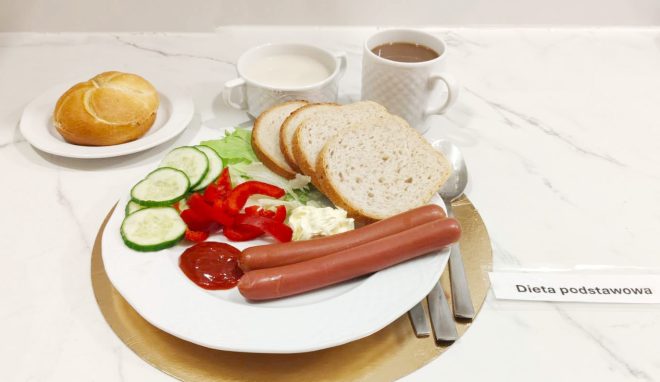 Śniadanie w diecie podstawowej podane na jasnym blacie. Na białym talerzu znajdują się dwie parówki, kromki pieczywa, plasterki ogórka i czerwonej papryki, sałata oraz porcja sosu i keczupu. Obok stoją kubki z mlekiem i kawą zbożową oraz bułka na osobnym talerzyku. Przy tacy ułożone są sztućce i kartka z napisem „Dieta podstawowa”