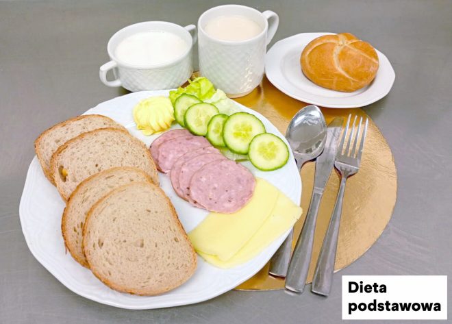 Śniadanie w diecie podstawowej na metalowym blacie. Na białym talerzu ułożono trzy kromki pieczywa, plastry ogórka i liście sałaty, porcję masła, kilka plastrów wędliny oraz plastry żółtego sera. W tle stoją dwa kubki z jasnymi napojami (np. mleko i kawa z mlekiem). Z prawej strony, na osobnym talerzyku, znajduje się bułka. Obok talerza ułożone są sztućce na okrągłej, złotej podkładce; w prawym dolnym rogu widoczny napis „Dieta podstawowa”