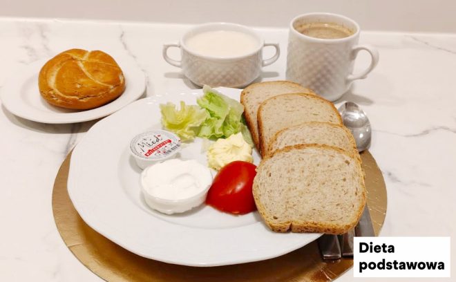 Śniadanie w diecie podstawowej podane na jasnym blacie. Na dużym białym talerzu leżą trzy kromki pieczywa pszennego, plaster pomidora, liście sałaty oraz porcja masła. Obok znajdują się dwa małe pojemniczki z dodatkami mlecznymi (np. jogurt/twarożek) oraz sztućce. W tle widać filiżankę kawy oraz miseczkę z białym napojem (np. mlekiem). Z lewej strony, na osobnym talerzyku, podana jest bułka. W prawym dolnym rogu widoczny napis „Dieta podstawowa”