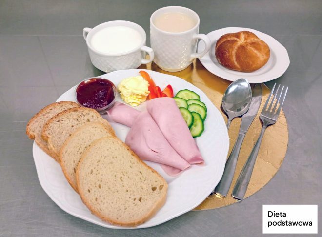Śniadanie na diecie podstawowej: kromki pieczywa pszennego, plasterki wędliny drobiowej, masło, warzywa świeże, porcja dżemu, bułka pszenna oraz kubki z mlekiem i napojem, podane na tacy