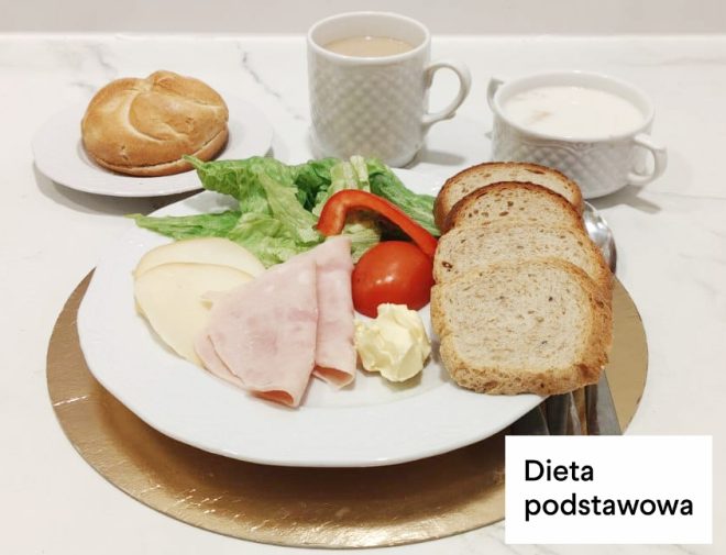 Śniadanie w diecie podstawowej: kanapki z chleba pszennego, wędlina drobiowa, ser żółty, masło, sałata i pomidor. Obok bułka pszenna oraz kubki z kawą zbożową i mlekiem. Na zdjęciu widoczny napis „Dieta podstawowa”