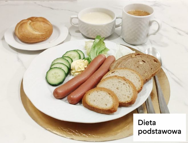 Śniadanie w diecie podstawowej: parówki podane z pieczywem pszennym, masłem, sałatą i plasterkami ogórka. Obok bułka pszenna oraz kubki z kawą zbożową i mlekiem. Na zdjęciu widoczny napis „Dieta podstawowa”