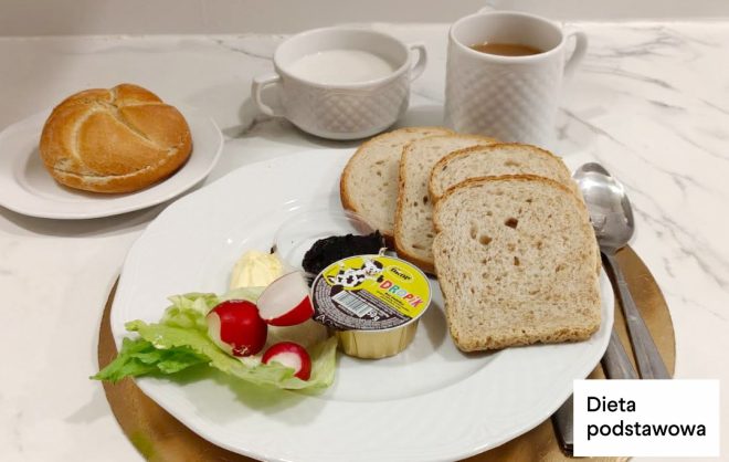 Śniadanie w diecie podstawowej: kanapki z chleba pszennego, twarożek, masło, sałata, rzodkiewka oraz porcja dżemu. Obok bułka pszenna oraz kubki z herbatą i mlekiem. Na zdjęciu widoczny napis „Dieta podstawowa”
