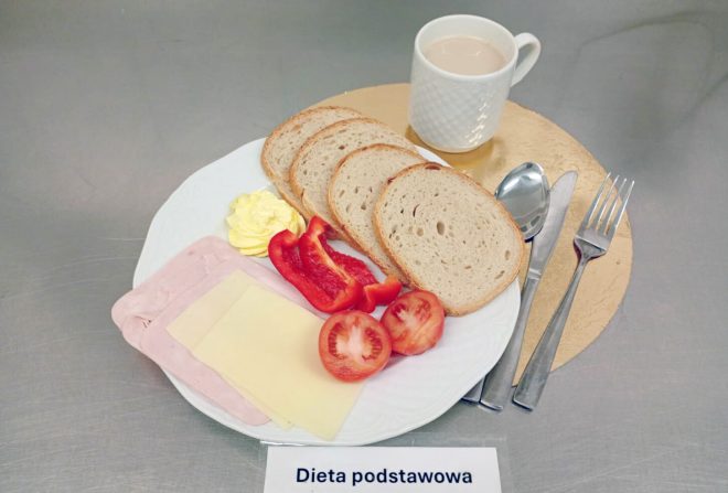 Śniadanie w diecie podstawowej: kromki chleba, plastry szynki drobiowej, plaster sera żółtego, masło, świeża papryka i pomidor. Obok kubek kawy z mlekiem. Widoczna etykieta „Dieta podstawowa”