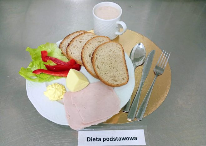 Śniadanie w „Diecie podstawowej” na metalowym blacie. Na białym talerzu ułożono trzy kromki pieczywa, plaster wędliny, kostkę sera, porcję masła, liść sałaty i paski czerwonej papryki. Z tyłu stoi biały kubek z jasnym napojem. Po prawej stronie leżą sztućce (łyżka, nóż i widelec) na złotej podkładce. Na dole kadru widoczna jest karteczka z napisem „Dieta podstawowa”