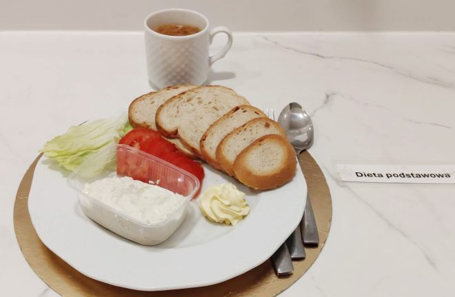 Śniadanie w „Diecie podstawowej” na jasnym, marmurowym blacie. Na białym talerzu leżą cztery kromki pieczywa, liść sałaty, plastry pomidora, a z przodu przezroczysty pojemnik z białym twarożkiem lub pastą. Obok znajduje się porcja masła w formie rozetki. Z tyłu stoi biały kubek z napojem (ciemniejszy, jak herbata). Po prawej stronie leżą sztućce na złotej podkładce, a obok widoczna jest karteczka „Dieta podstawowa”