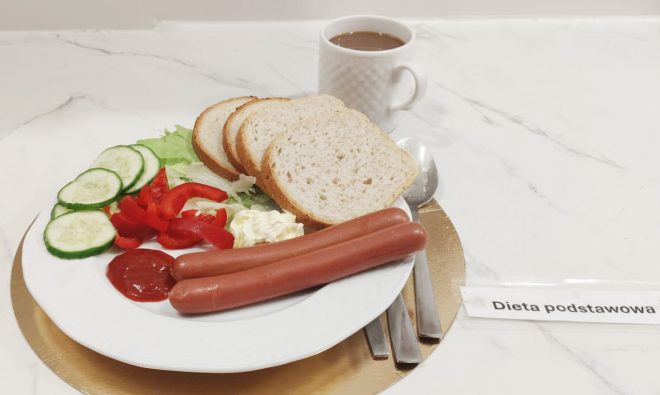Śniadanie „Dieta podstawowa” na jasnym, marmurkowym blacie: na białym talerzu dwie parówki, kilka kromek białego chleba, plasterki ogórka i czerwonej papryki na liściach sałaty oraz porcja majonezu/sosu i ketchupu. W tle stoi biały kubek z kawą lub herbatą z mlekiem, obok leżą sztućce. Z prawej strony widoczna kartka z napisem „Dieta podstawowa”