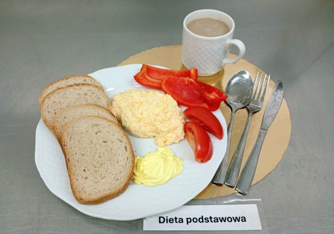 Śniadanie „Dieta podstawowa” na metalowym blacie: na białym talerzu trzy kromki chleba, porcja pasty/jajecznej sałatki, plasterki czerwonej papryki i cząstki pomidora oraz porcja żółtego masła/margaryny. Z prawej strony ułożone są sztućce, a w tle stoi biały kubek z jasnym napojem (kawa lub herbata z mlekiem). U dołu kadru widoczna kartka z napisem „Dieta podstawowa”
