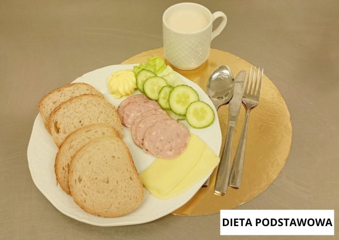 Śniadanie w diecie podstawowej: na białym talerzu kromki chleba, plastry wędliny i sera żółtego, masło, ogórek świeży i liść sałaty; obok filiżanka mleka oraz sztućce