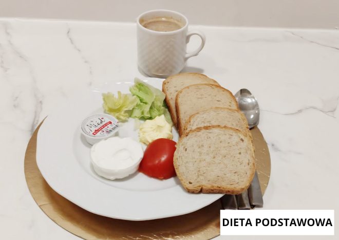 Śniadanie w diecie podstawowej podane na białym talerzu. Na talerzu znajdują się kromki chleba pszennego, porcja twarożku, sałata, plaster pomidora oraz kawałek masła. Obok ustawiona jest filiżanka kawy. Zestaw ułożony na jasnym blacie, z widocznym napisem „Dieta podstawowa”