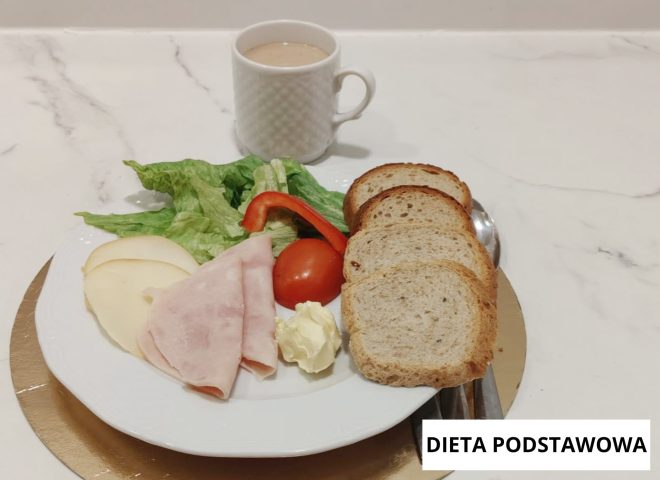 Śniadanie w diecie podstawowej: pieczywo pszenne, wędlina drobiowa, ser żółty, masło, sałata, pomidor i papryka. Obok kubek z kawą zbożową. Na zdjęciu widoczny napis „Dieta podstawowa”