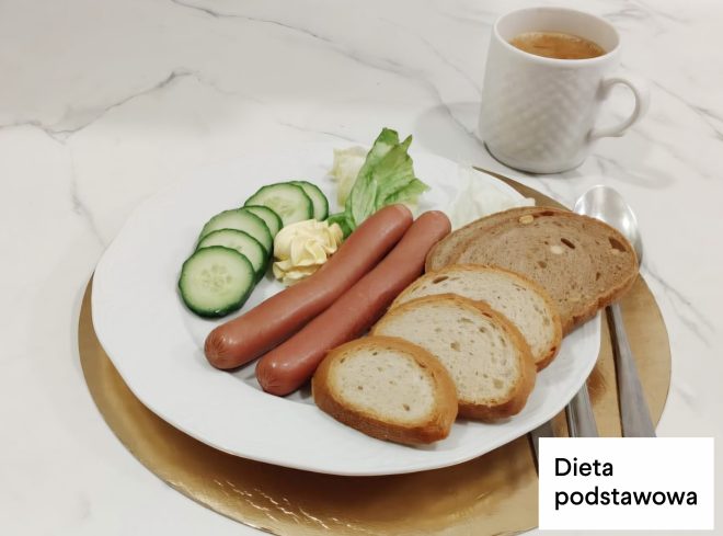 Śniadanie w diecie podstawowej: parówki podane z pieczywem pszennym, masłem, sałatą i plasterkami ogórka. Obok kubek z kawą zbożową. Na zdjęciu widoczny napis „Dieta podstawowa”