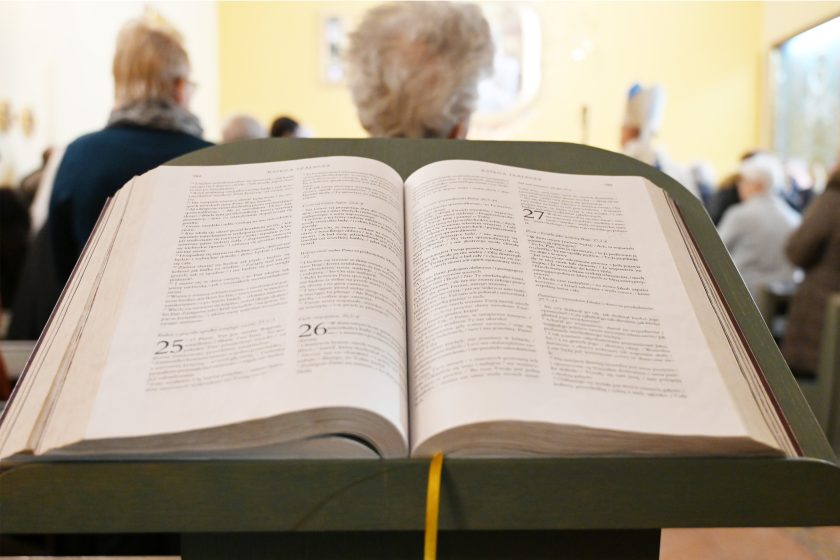 Otwarte Pismo Święte leżące na pulpicie w kaplicy; na pierwszym planie widoczne strony księgi, w tle rozmyte sylwetki wiernych stojących w ławkach