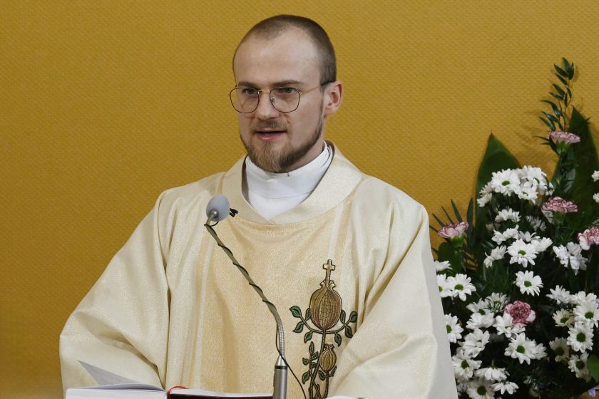 Brat zakonny w jasnym ornacie z haftowanym symbolem zakonu stoi przy ambonie i czyta z otwartej księgi podczas Mszy Świętej; obok mikrofon, w tle żółta ściana i kompozycja z białych kwiatów