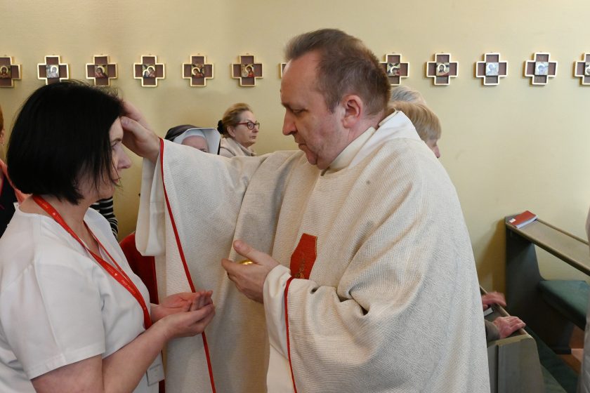 Kapłan w jasnym ornacie udziela błogosławieństwa kobiecie w stroju medycznym z identyfikatorem; duchowny unosi dłoń nad jej głową, a kobieta trzyma ręce złożone w geście modlitwy