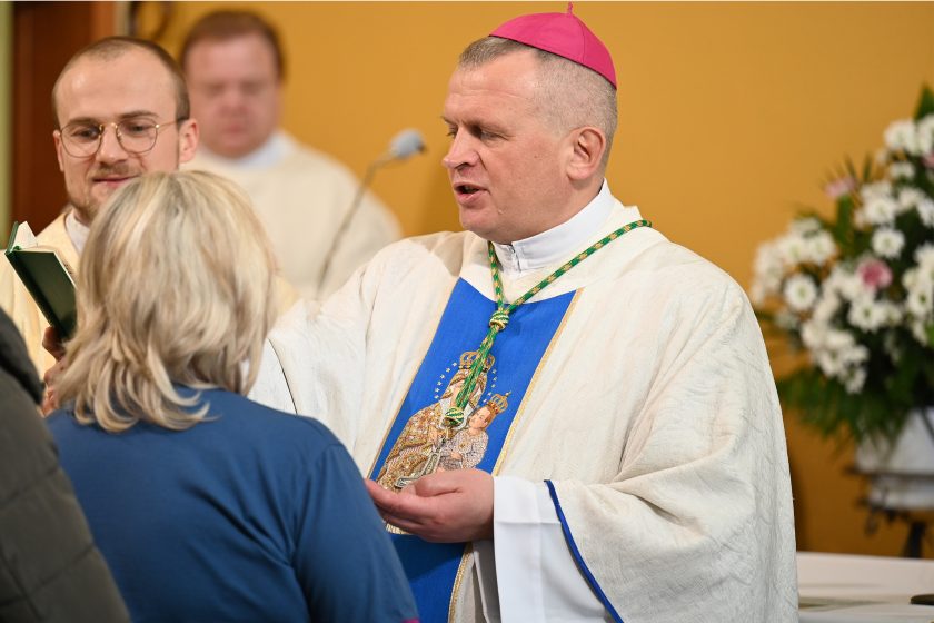 Biskup w białych szatach liturgicznych z niebieskim pasem i różową piuską udziela sakramentu wiernej kobiecie; obok stoi brat zakonny trzymający otwartą księgę liturgiczną