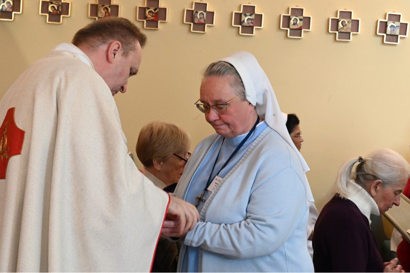 Kapłan w jasnym ornacie pochyla się nad siostrą zakonną w niebieskim habicie i białym welonie, udzielając jej Komunii lub błogosławieństwa; w tle widoczni inni uczestnicy liturgii