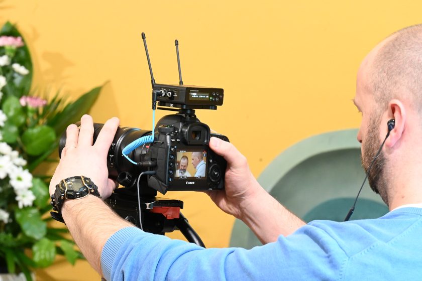 Mężczyzna obsługujący kamerę filmową nagrywa przebieg uroczystości w kaplicy; na ekranie aparatu widoczna scena liturgiczna z duchownymi, w tle żółta ściana i kwiaty