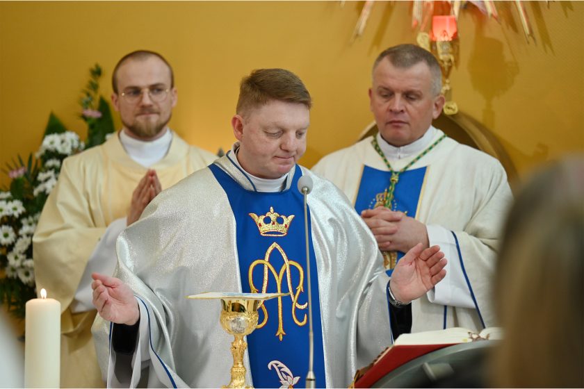 Kapłan w niebiesko-srebrnych szatach liturgicznych modli się przy ołtarzu z rozłożonymi rękami; w tle biskup oraz brat zakonny stoją w skupieniu podczas Eucharystii