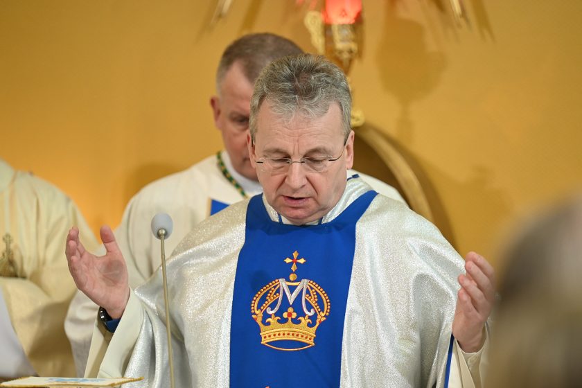 Zbliżenie na kapłana w srebrno-niebieskim ornacie z haftowaną koroną i symbolem maryjnym; duchowny unosi ręce w geście modlitwy podczas liturgii, w tle widoczny biskup