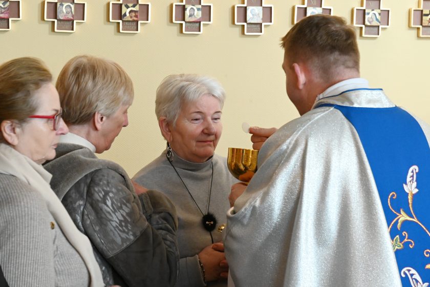 Kapłan udziela Komunii Świętej kobiecie stojącej w kolejce; wierna patrzy z skupieniem, a duchowny trzyma Hostię i kielich. Obok stoją inne kobiety oczekujące na przyjęcie sakramentu