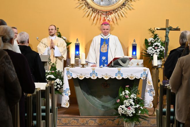 Biskup w białych szatach liturgicznych z niebieskim pasem stoi przy ołtarzu podczas Eucharystii; obok niego brat zakonny ze złożonymi dłońmi. Na ołtarzu świece i kwiaty, nad nim dekoracyjny element sakralny