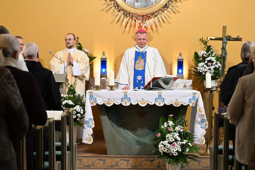 Biskup w białych szatach liturgicznych z niebieskim pasem stoi przy ołtarzu podczas Eucharystii; obok niego brat zakonny ze złożonymi dłońmi. Na ołtarzu świece i kwiaty, nad nim dekoracyjny element sakralny