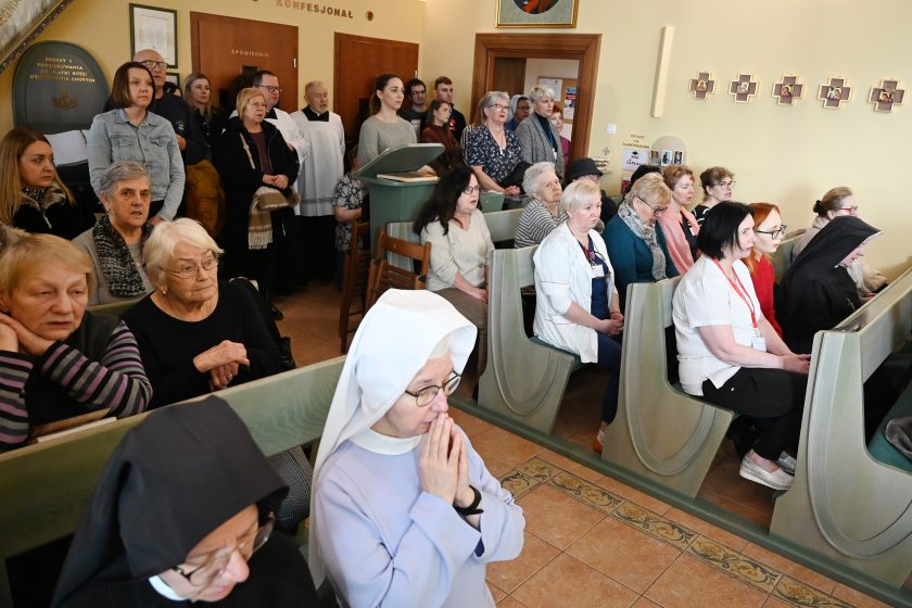 Wnętrze kaplicy szpitalnej wypełnione wiernymi – kobiety i mężczyźni stoją oraz siedzą w ławkach podczas liturgii; na pierwszym planie widoczna siostra zakonna z rękami złożonymi do modlitwy, w tle personel medyczny w białych strojach