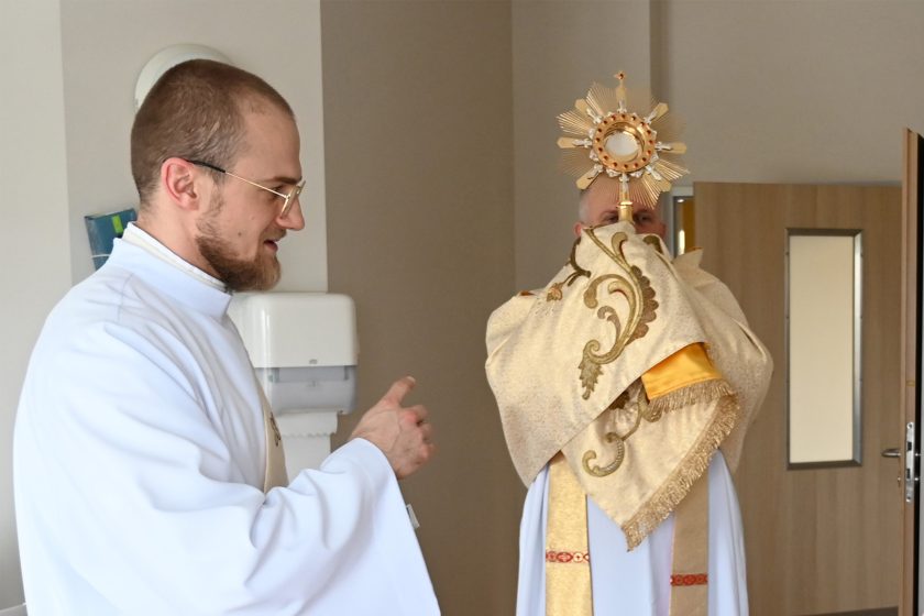 Brat zakonny w jasnym habicie wskazuje drogę lub udziela wskazówek kapłanowi niosącemu monstrancję z Najświętszym Sakramentem na szpitalnym korytarzu. W tle widoczne drzwi do sal oraz elementy wyposażenia medycznego