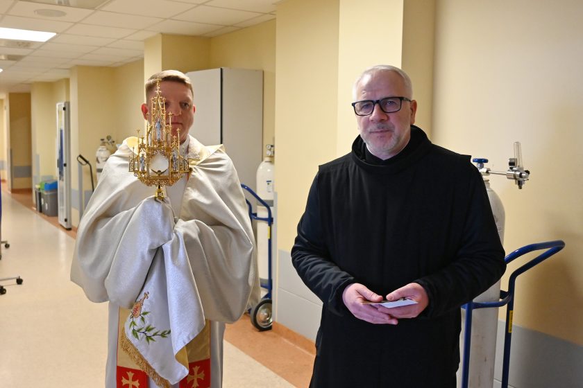 Dwóch braci zakonnych stoi na szpitalnym korytarzu. Jeden z nich w jasnym ornacie trzyma bogato zdobioną monstrancję z Najświętszym Sakramentem, drugi w czarnym habicie trzyma w dłoniach obrazki religijne. W tle widoczne są drzwi do sal chorych oraz sprzęt medyczny