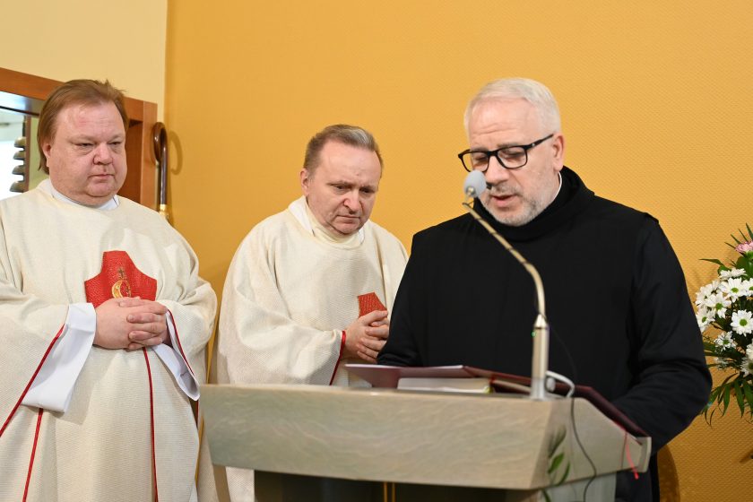 Trzech duchownych przy ambonie – jeden czyta tekst z księgi, dwaj pozostali stoją obok ze złożonymi rękami w geście modlitwy