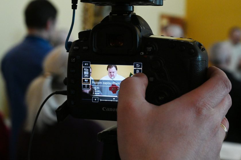 Zbliżenie na ekran aparatu fotograficznego rejestrującego przebieg Mszy Świętej; na wyświetlaczu widoczny duchowny przy ambonie, w tle rozmyte postacie uczestników liturgii